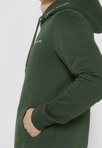 Olive green hoodie with a drawstring hood, kangaroo pocket, and cuffed sleeves. Features "TOMMY HILFIGER" logo and "EST.1985" text.