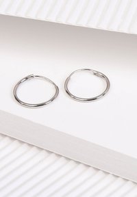 Silberne Creolen mit einer glatten, polierten Oberfläche, kreisförmiger Form und sicherem Schnappverschluss, die auf einem weißen strukturierten Hintergrund liegen.