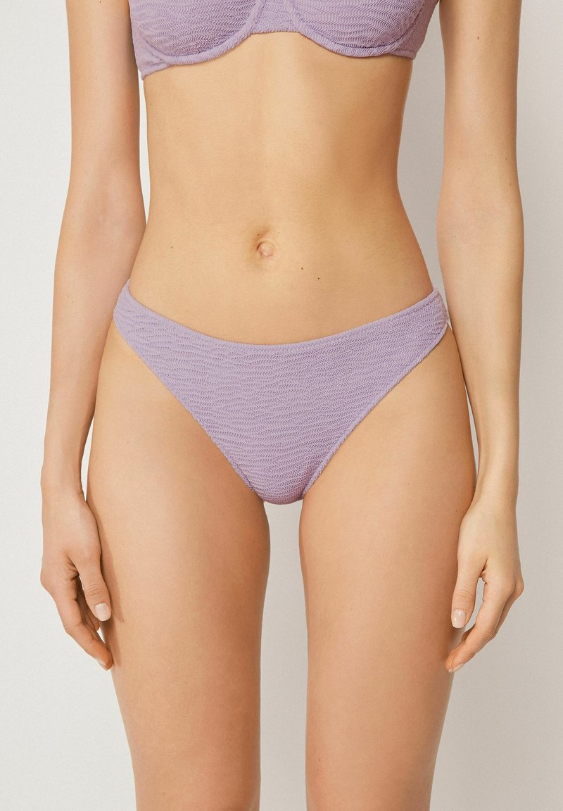 Frau trägt strukturierte hellviolette Bikinihose und passendes Oberteil, steht vor einem einfarbigen hellen Hintergrund.