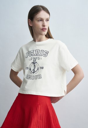 Žena v bielom krátkom tričku s nápisom "Paris Amour 1998" a grafikou kotvy, v červenej plisovanej sukni, stojaca pred svetlým pozadím.