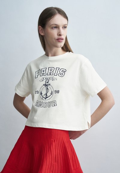Γυναίκα που φοράει λευκό κοντό T-shirt με την επιγραφή "Paris Amour 1998" και γραφικό άγκυρας, συνδυασμένο με κόκκινη πλισέ φούστα, στέκεται μπροστά σε ανοιχτό φόντο.