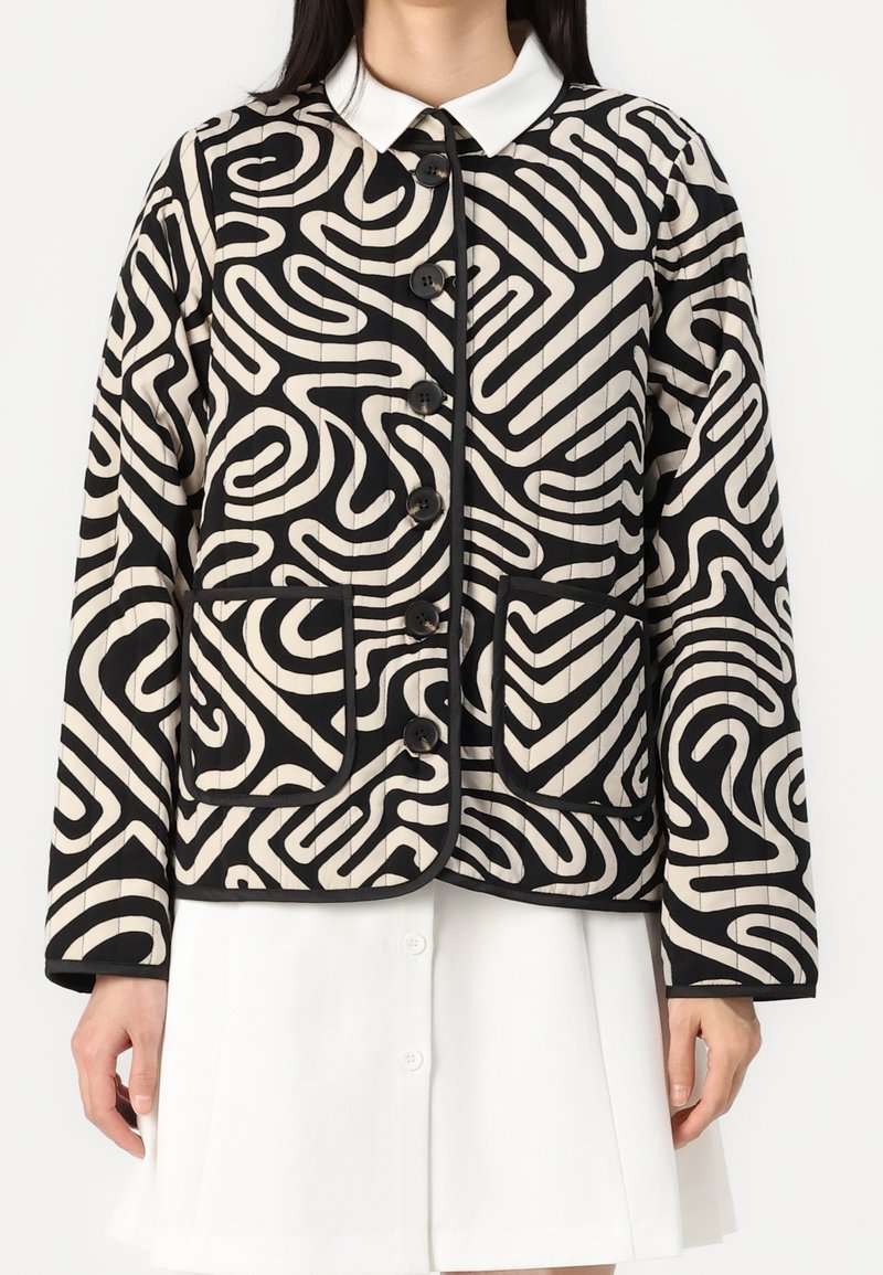 Femme portant une veste boutonnée à motif noir et blanc avec deux poches avant, par-dessus une jupe blanche.