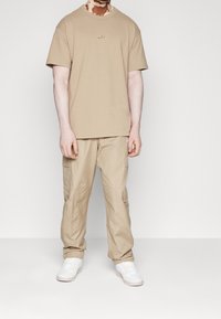 Beige tričko s krátkým rukávem a malým logem, spárováno s béžovými cargo kalhotami s kapsami a bílými teniskami. Jednoduchý, ležérní design.