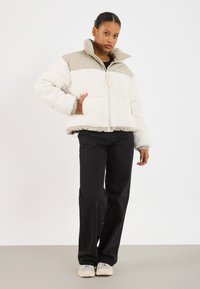 Teksturowana kremowo-beżowa kurtka z wysokim kołnierzem, zamkiem błyskawicznym, bocznymi kieszeniami i elastycznymi mankietami, zestawiona z czarnymi spodniami i sneakerami.