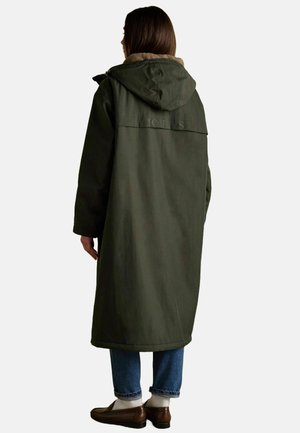 Langer grüner Mantel mit Kapuze, aus strukturiertem Stoff. Hat ein hellbraunes Innenfutter und ein geprägtes Logo auf dem Rücken. Getragen über Jeans und Schuhen.