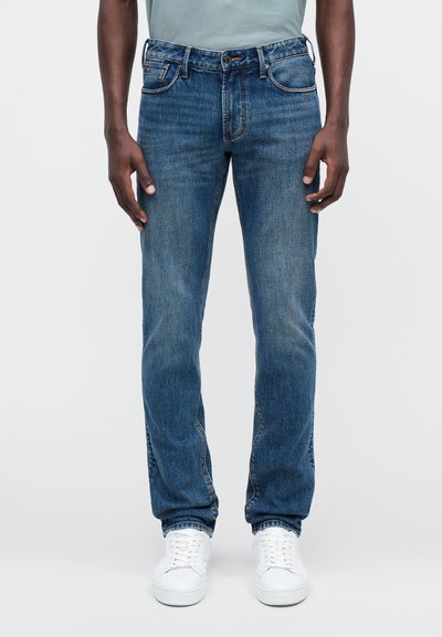 Μπλε τζιν παντελόνι με ίσια γραμμή, ελαφριά φθορά, σχέδιο πέντε τσεπών και αντίθετη ραφή, συνδυασμένο με λευκά αθλητικά παπούτσια.