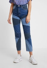 Žena v modrých patchwork džínách s různými odstíny, fialové vzorované blůze a bílých teniskách, stojící před jednobarevným šedým pozadím.