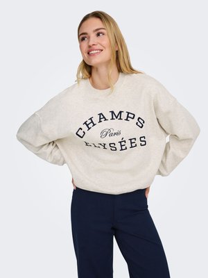 Lachende vrouw met lang blond haar, gekleed in een beige sweatshirt met de tekst "CHAMPS Paris ELYSÉES" en donkerblauwe broek, handen in de zij.