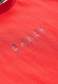 Camiseta de algodón roja con un logo en relieve de color plateado metálico que dice "BAKER by TED BAKER." Diseño simple con cuello redondo y textura suave.