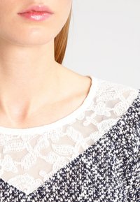 Stickad tröja med vit spets i axlarna, med marinblå och vit textur mönster, rund halsringning och mjuk tygstruktur.