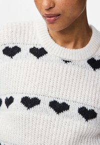 Vit stickad tröja med svarta hjärtan, rund hals och ribbat avslut, med ett mjukt, texturerat tyg.