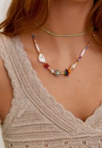 Collier coloré composé de perles mélangées incluant des perles, du verre et des coquillages, posé sur un haut en maille beige texturé avec un décolleté en V profond.