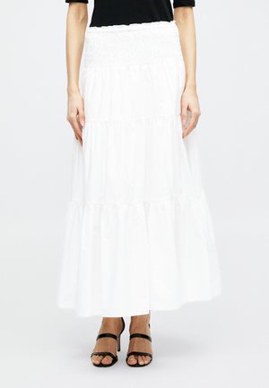 Γυναίκα που φορά μακριά λευκή φούστα με στρώσεις, μαύρο τοπ και μαύρα σανδάλια με λεπτά λουράκια και ψηλό τακούνι, σε απλό φόντο.