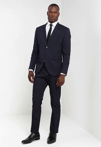 Laivastonsininen puku, jossa on yksirivinen malli, lovellut kaulukset ja kaksi taskua. Yhdistetty valkoiseen paitaan, mustaan solmioon ja kiiltäviin mustiin kenkiin.