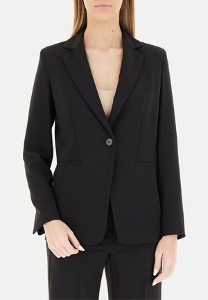 Donna che indossa un blazer nero aderente con un bottone sopra una maglia beige, abbinato a pantaloni neri coordinati, in piedi davanti a uno sfondo semplice.