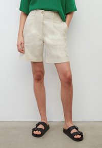 Beige puuvillashortsit, joissa on laskokset, etutaskut ja suora lahje. Yhdistetty mustiin sandaaleihin, joissa on soljellinen yksityiskohta.
