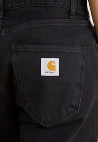 Svarte denimbukser har en baklomme med en hvit etikett som viser en gul logo og teksten "carhartt." Teksturen er jevn og solid.