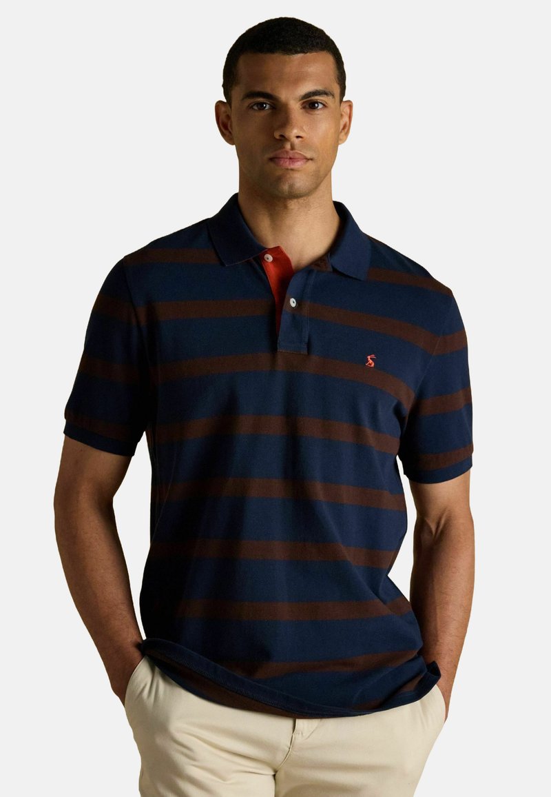 Jūras zils polo krekls ar brūnām horizontālām svītrām, ar sarkanu apkakli un nelielu logo uz krūtīm. Īsās piedurknes, kokvilnas audums.