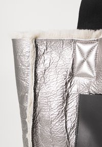 Hõbedane metallik kott tekstuurse viimistlusega, millel on pehme kreemivillane vooder ja mustad detailid, sealhulgas tikkimiseks mõeldud detail.