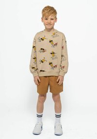 Felpa beige con illustrazioni di cartoni animati, collo rotondo e maniche lunghe abbinata a pantaloni corti marroni. Il modello indossa sneaker bianche con accenti blu.
