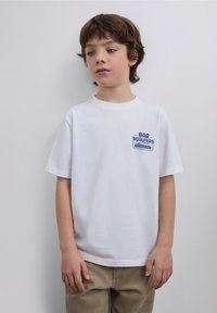 White cotton t-shirt featuring a blue printed logo on the left chest. The design is simple with short sleeves and a crew neckline.