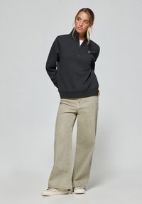 Svart zip-up sweatshirt med en liten logo på vänster bröst, kombinerad med ljusbeiga vida byxor och vita sneakers.