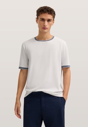 Junger Mann mit lockigem Haar, der ein weißes Kurzarm-T-Shirt mit dunklem Rand und dunkelblaue Hose trägt, steht mit einer Hand in der Tasche.