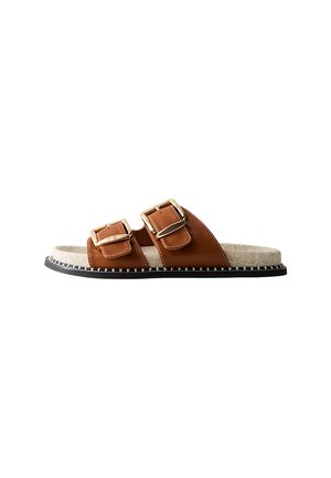 Bruna fusk läder slide sandal med två justerbara gyllene spännen, en beige mjuk fotbädd och en svart yttersula med vit sömnadsdetalj.