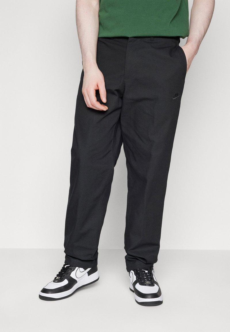 Pantalon noir à coupe décontractée, arborant un logo Nike subtil. Associé à des baskets noir et blanc, ainsi qu'une chemise à manches courtes verte.
