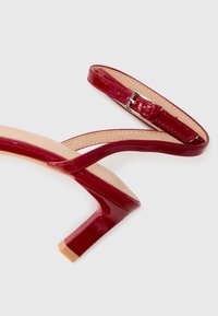 Red patent leather sandal with a rounded toe, thin straps, and an ankle buckle. Features a block heel and a smooth beige insole.