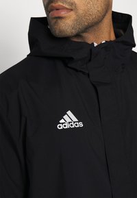 Czarna kurtka z wysokim kołnierzem i kapturem, wykonana z gładkiego materiału. Posiada białe logo Adidas na lewej piersi.