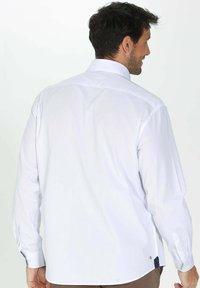 Chemise blanche à manches longues avec un col boutonné, présentant une texture lisse et des accents de poignet contrastants. Le design inclut une petite étiquette de logo.