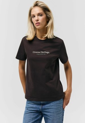 Kobieta o blond włosach do ramion, ubrana w czarną koszulkę z napisem "Różnorodne Dziedzictwo" i niebieskie dżinsy, stojąca na tle jasnego, gładkiego tła.