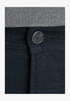 Tmavě modrá denímová tkanina s šedým pleteným pasem. Obsahuje černý kovový knoflík s vyrytým vzorem a pevné stehy podél švu.