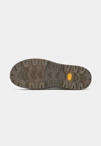 Såle av brun fotturssko med dype, flerretningede dekkmønstre og en gul Vibram-logo nær buen.
