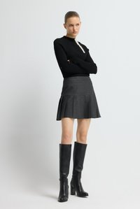 Zwart gebreid shirt met lange mouwen en een gestructureerde knoopdetail, gecombineerd met een grijze geplooide minirok en zwarte leren laarzen met hak tot aan de knie.