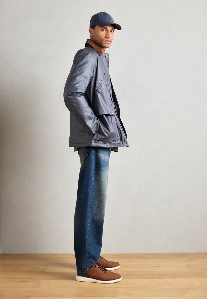 Veste grise isolante avec un col marron, associée à un jean en denim bleu et des chaussures marron. Le modèle se tient de profil devant un fond neutre.