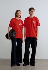 Κόκκινα βαμβακερά t-shirts με το λογότυπο «Calvin Klein»; συνδυασμένα με σκούρα τζιν παντελόνια καμπάνα. Ένα μοντέλο κρατά μια μαύρη δερμάτινη τσάντα. Απλός σχεδιασμός.