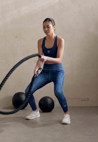 Ropa deportiva de patrón azul, que incluye una camiseta sin mangas y leggings, con una mujer agarrando una cuerda de batalla negra. Hay dos bolas de ejercicio negras cerca.