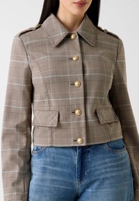 Veste courte à carreaux marron avec boutons dorés, col pointu et deux poches avant. Le tissu présente une finition texturée avec des accents bleus.