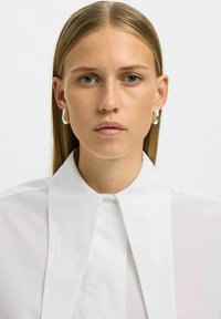 Noor inimene sirgete blondide juustega, kandes hõbedasi rõngaste kõrvarõngaid ja valget särki laia kraega, vaatab otse vastu ühtlase tausta vastu.