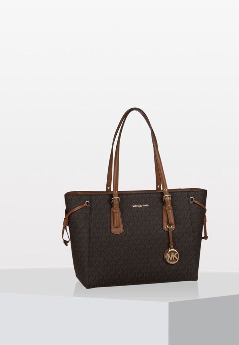Sac cabas Michael Kors marron foncé avec motif monogramme, deux longues poignées en cuir et un charm logo métallique circulaire suspendu à l'avant.
