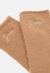 Bracciali per le braccia in maglia di colore tan con una texture morbida; presenta il logo "Guess" ricamato in filo metallico su un lato. I bordi sono leggermente sfrangiati.