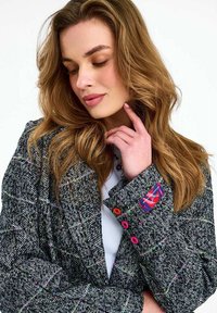 Žena so zvlnenými svetlohnedými vlasmi nosí čierno-biely sako s farebnými gombíkmi a červenou štítkovou značkou, pričom si zamyslene dotýka brady.