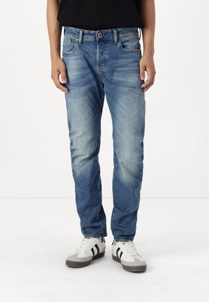 Person iført blå falmede jeans og hvide sneakers med sorte striber, stående mod en ensfarvet hvid baggrund.