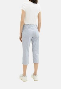 Donna che indossa pantaloni blu chiaro a vita corta, una maglietta bianca a maniche corte e scarpe da ginnastica bianche, in piedi di spalle su uno sfondo semplice.