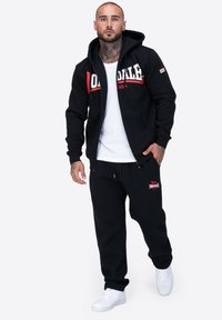 Czarna bluza z kapturem na zamek z czerwonym i białym logo, połączona z czarnymi spodniami dresowymi, z haftem. Białe trampki dopełniają stylizację.