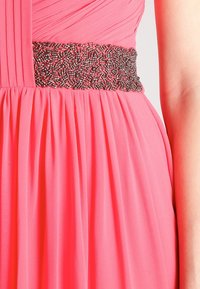Abito rosa a pieghe con una vita accentuata in argento decorata con perline. Il tessuto sembra leggero e dalla consistenza morbida, creando una silhouette fluida.