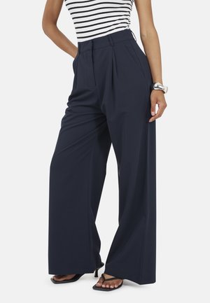 Persoon die marineblauwe, wijde broek met hoge taille draagt, zwarte sandalen met hak en vierkante neus, gestreept mouwloos topje en zilveren horloge aan linkerpols.