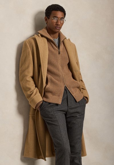 Bărbat purtând o haină de lână bej, pulover maro cu fermoar tricotat, pantaloni gri și ochelari, stând sprijinit de un perete bej cu mâinile în buzunare.